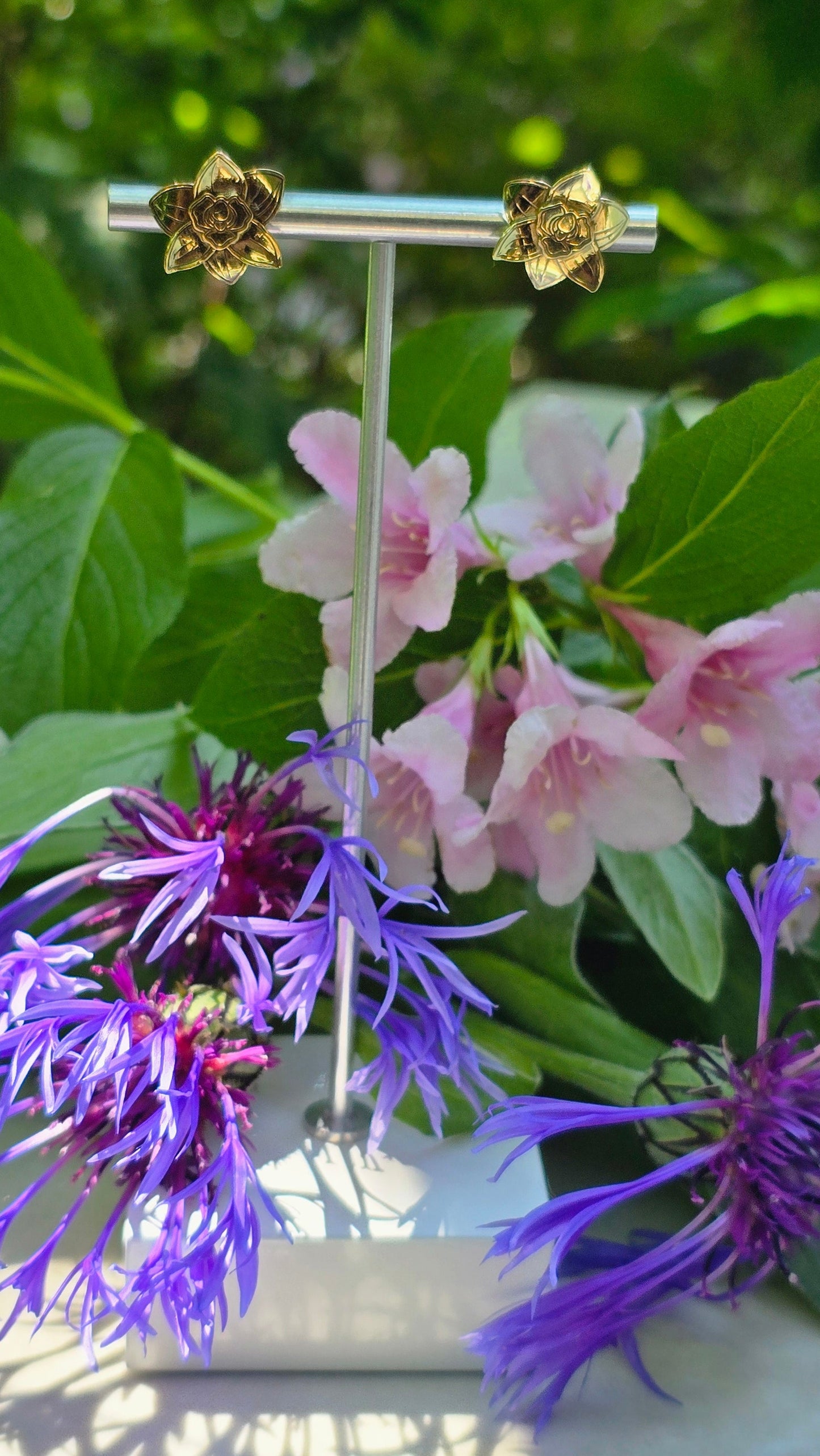 Gold flower-shaped earrings on a stand with purple and pink flowers in the background