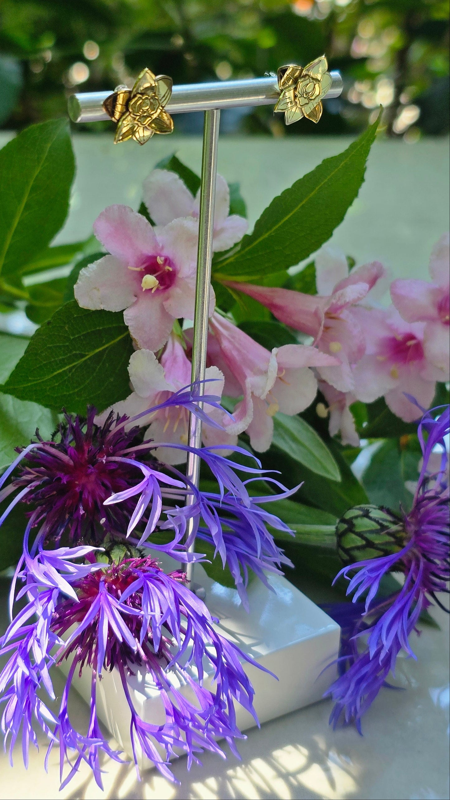 Gold flower-shaped earrings on a stand with pink and purple flowers in the background