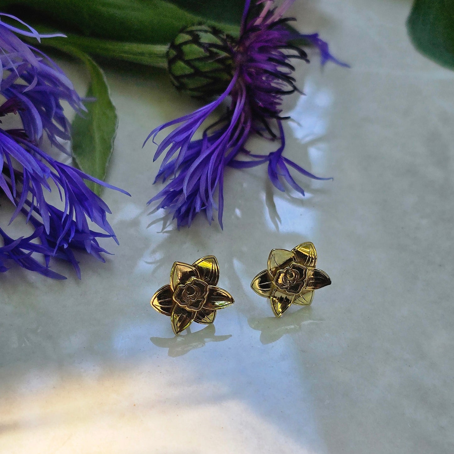Gold flower-shaped earrings on a white surface with purple flowers in the background