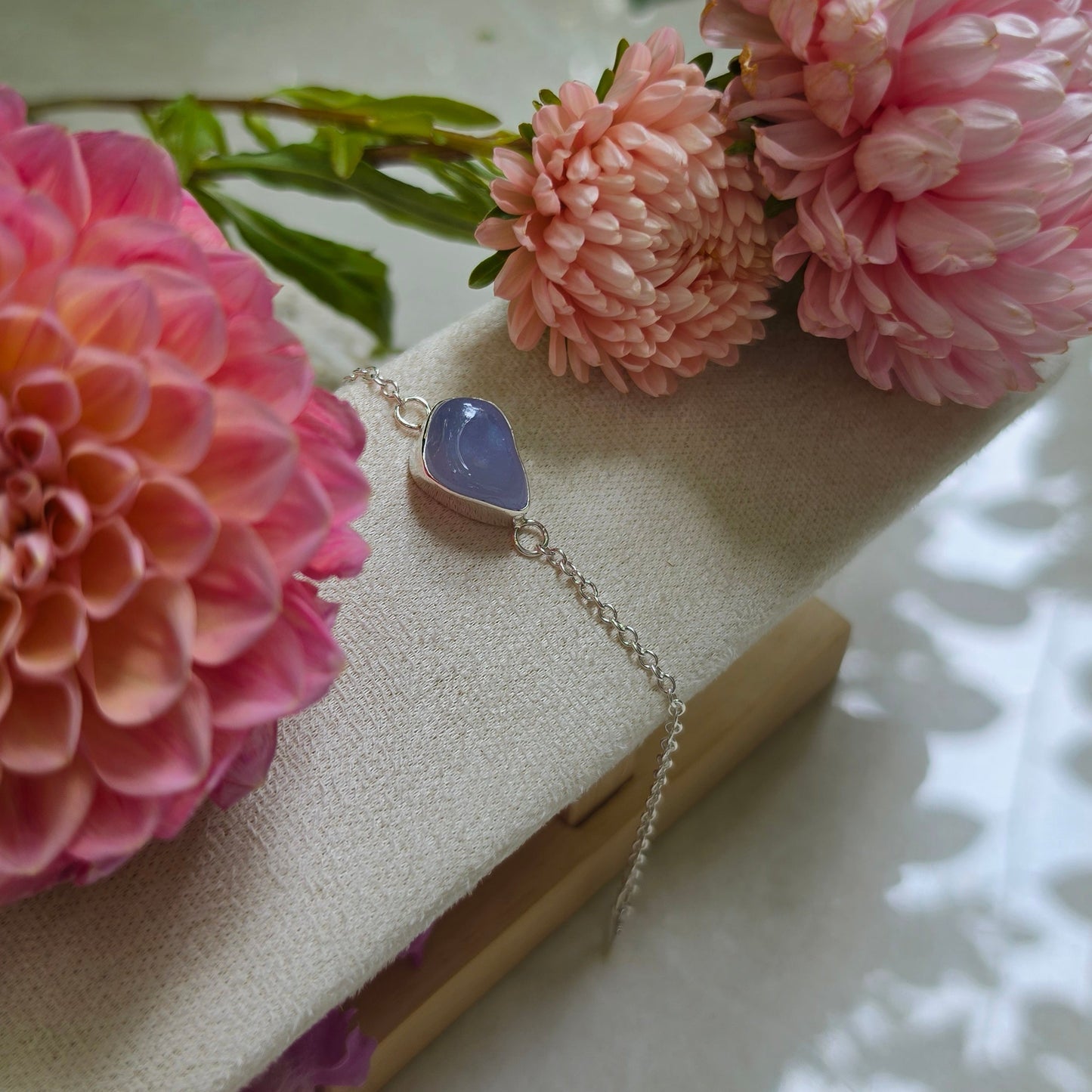 A delicate silver chain bracelet with a small oval light blue stone pendant, styled on a cream-colored jewelry display block surrounded by pink flowers.