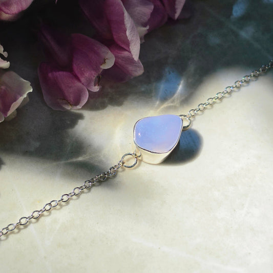 A delicate silver chain bracelet featuring a small, polished Blue Chalcedony stone pendant, displayed on a textured beige surface with purple flowers in the background.