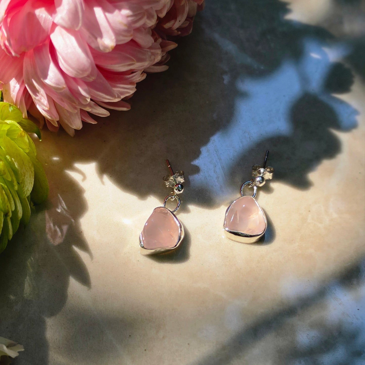 A pair of silver-toned dangle earrings with irregular light pink stone drops, styled on a sunlit beige surface next to a pink flower.