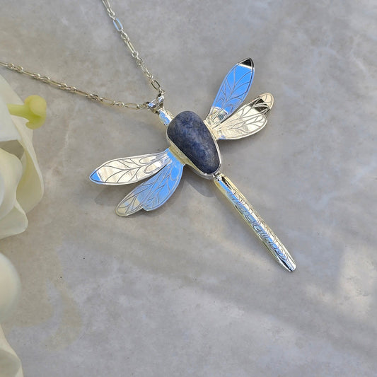 Sterling silver dragonfly necklace featuring a bezel-set dumortierite stone forming the body, with intricately laser-engraved wings and tail for fine detail. Suspended on a chain-link style sterling silver chain, showcasing deep blue tones and delicate craftsmanship.